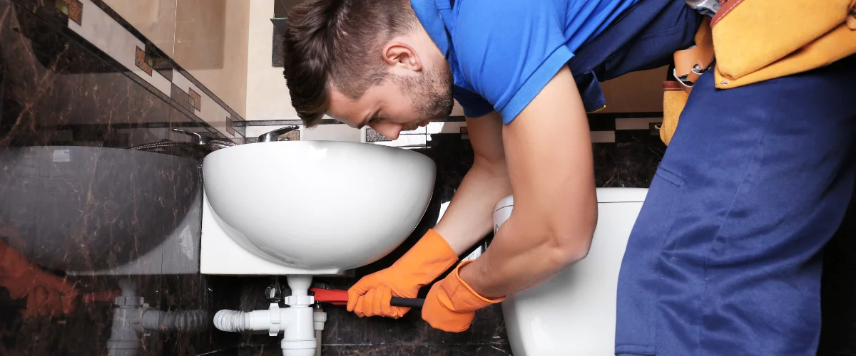 A repairman in a green sweater using a wrench to fix the chrome P-trap and pipes under a sink.