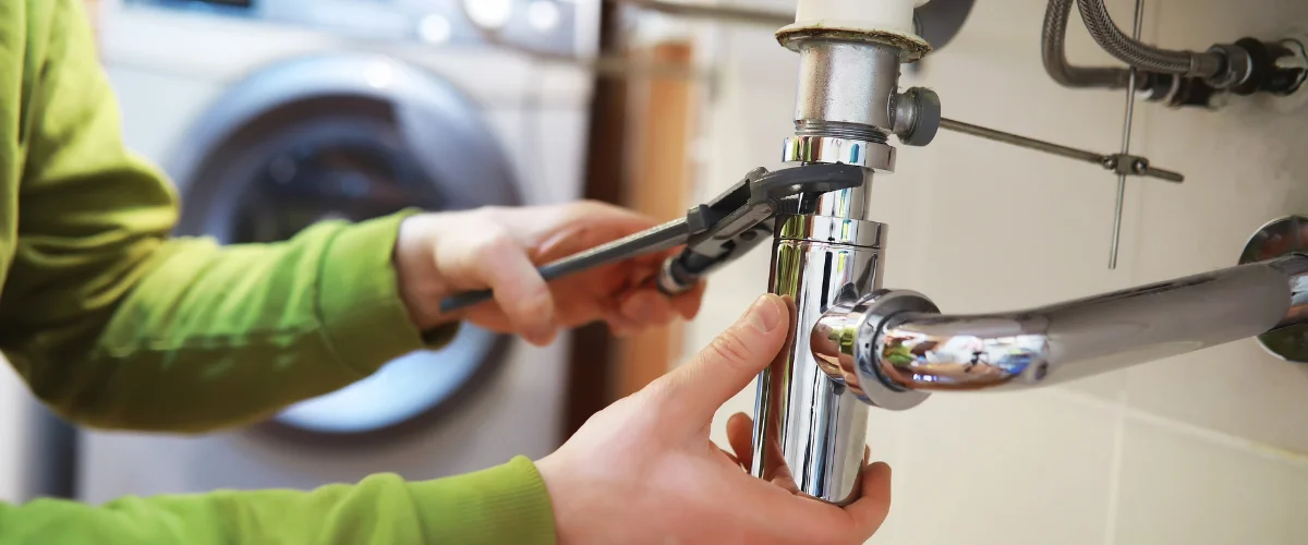 A male plumber in blue uniform and orange gloves fixing a white bathroom sink with a wrench.