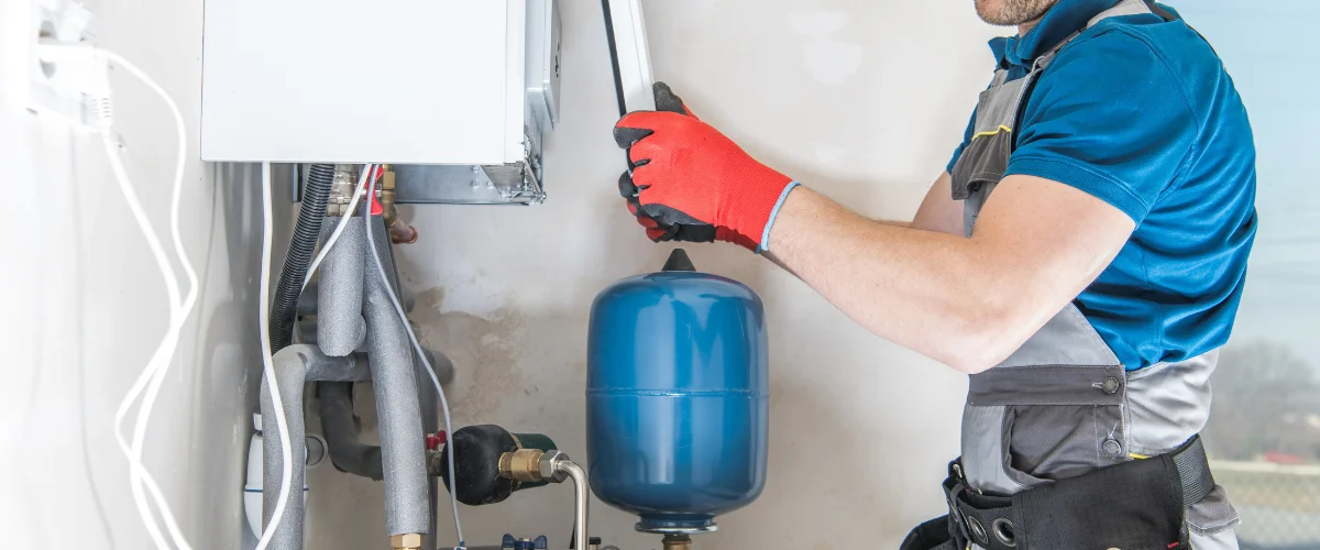 A gloved plumber in blue and grey work clothes installing or maintaining a white boiler and blue expansion tank. A gloved plumber in blue and grey work clothes installing or maintaining a white boiler and blue expansion tank.