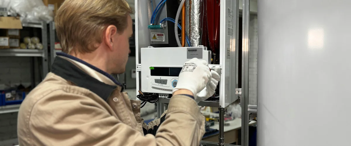 A technician wearing a tan jacket and white gloves works on fixing a white heating appliance in a workshop setting.