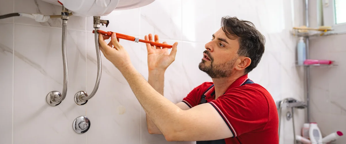 A construction worker in an orange hard hat and blue overalls installs red and blue plumbing pipes inside a building.