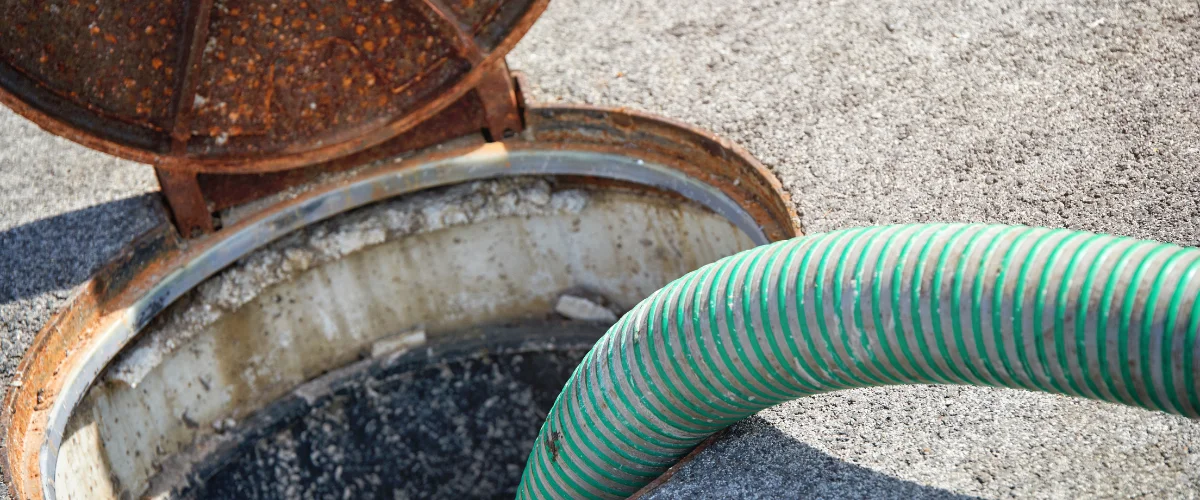 A green and white ribbed hose extends into an open, rusty manhole cover of a septic tank.