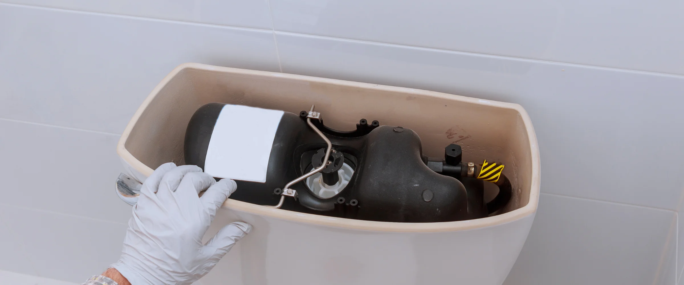 A gloved hand inspecting the complex black internal mechanism of an open toilet tank against a white tiled wall.