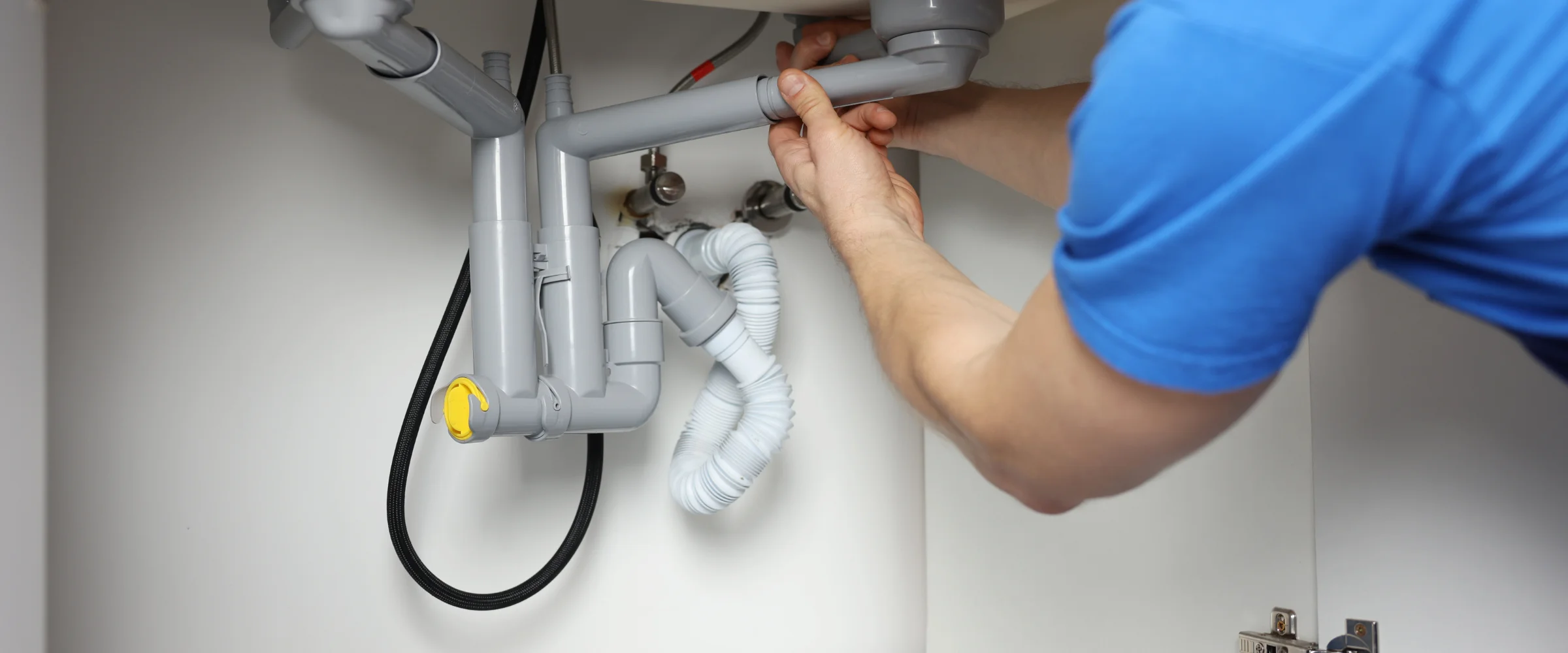 A plumber in a blue shirt installing or repairing grey plastic drainage pipes under a sink in a white cabinet.