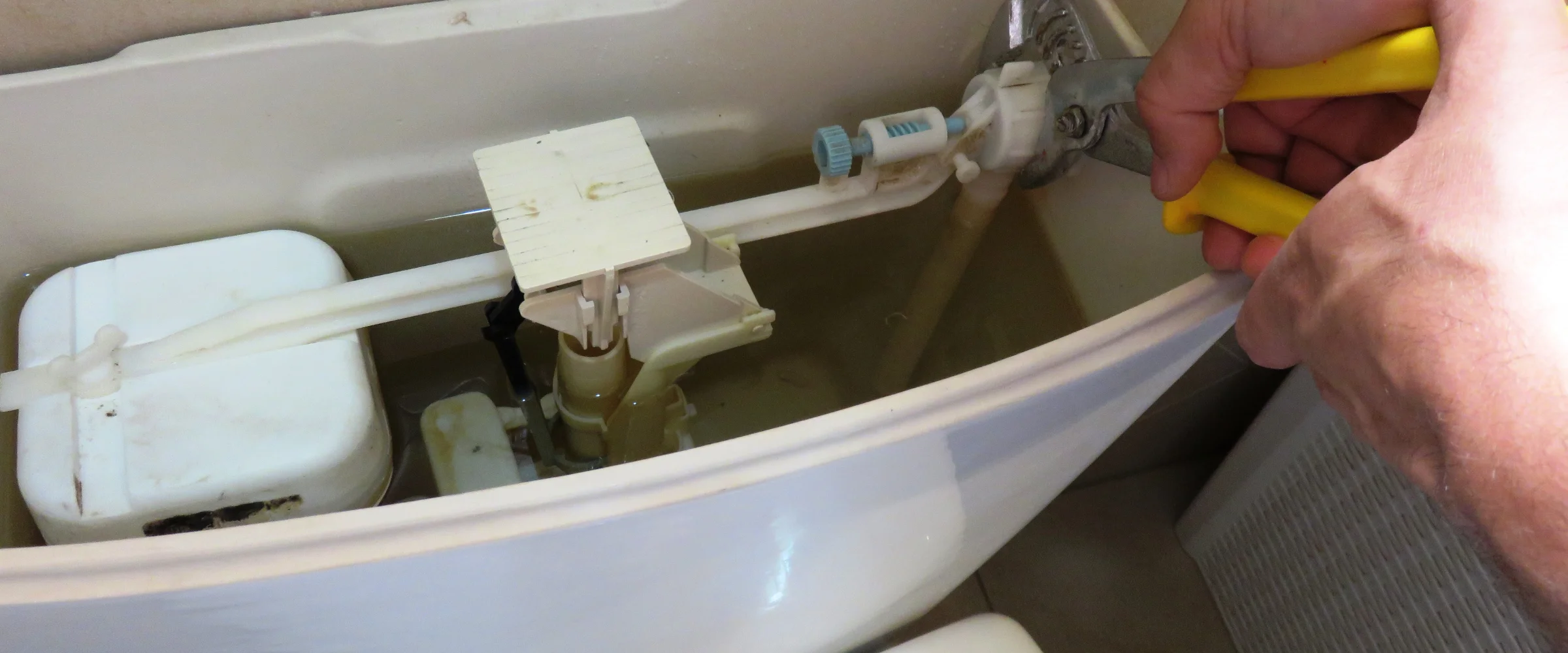 A close-up view of a person's hand using pliers to adjust the fill valve inside a toilet tank.