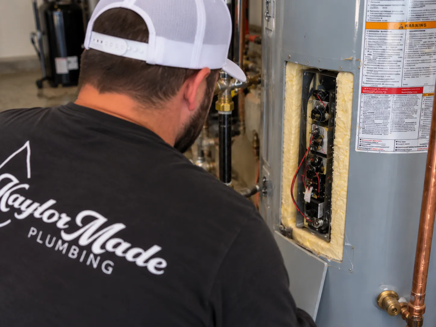 A plumber, seen from the back, works on the exposed components of a water heater with insulation and wiring visible.