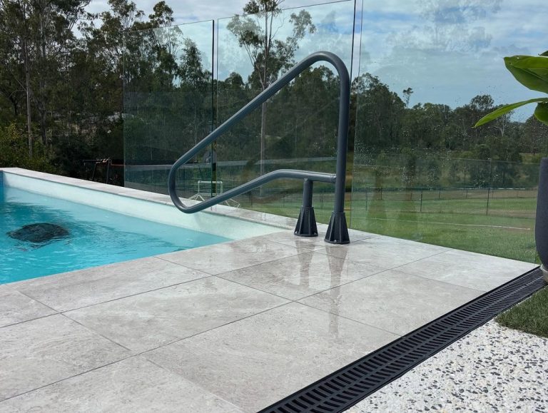 A black metal pool safety hand rail installed on a paved pool edge beside glass fencing