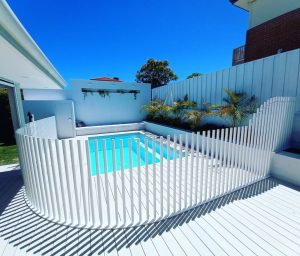A backyard swimming pool enclosed by a white picket-style pool fence under blue sky
