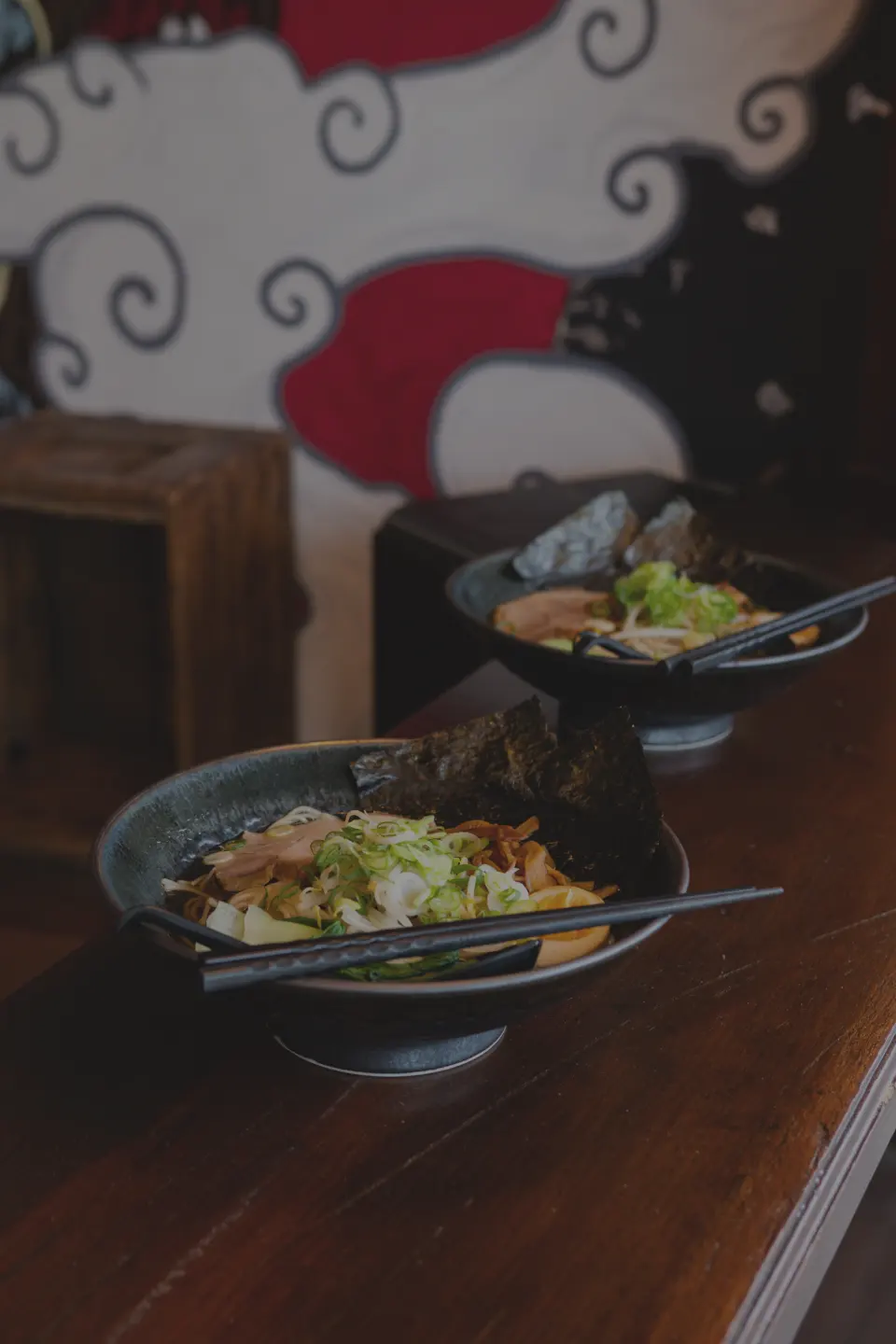 Two bowls of ramen with nori and chopsticks on a timber counter below the mural