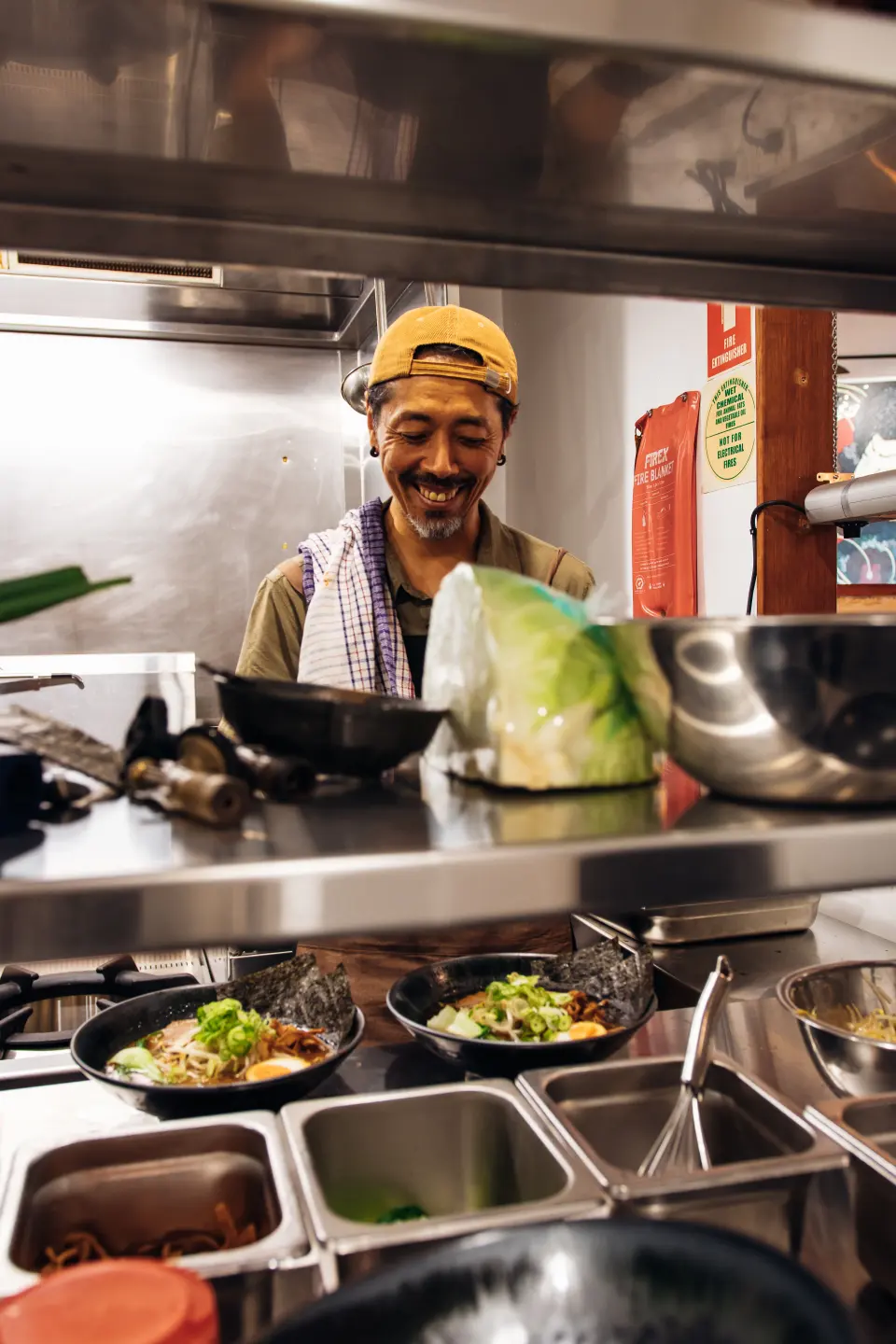 A smiling chef in a yellow cap behind the pass as bowls of ramen are finished