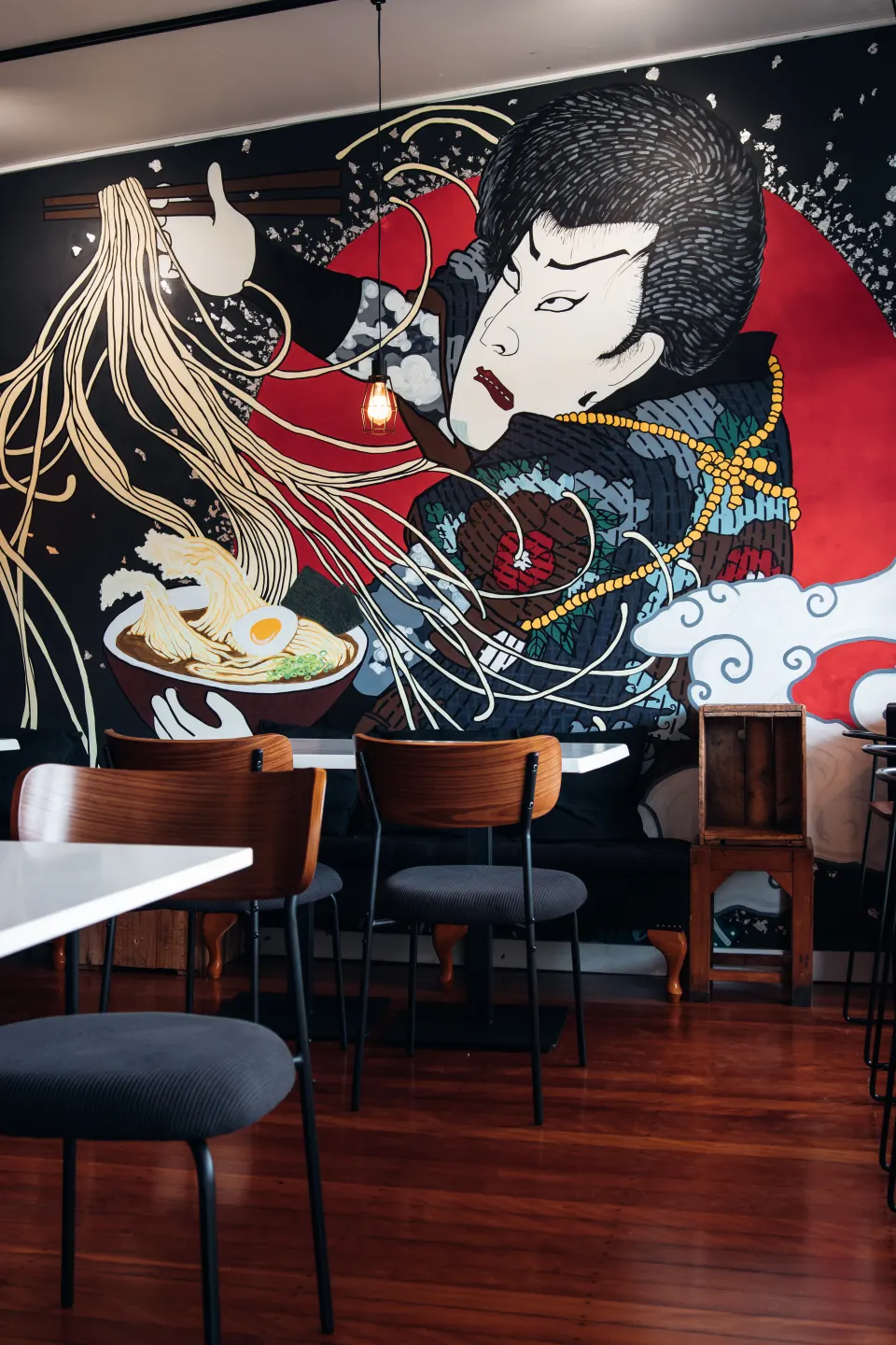 Wooden tables and chairs below the Japanese ramen mural at Pompoko
