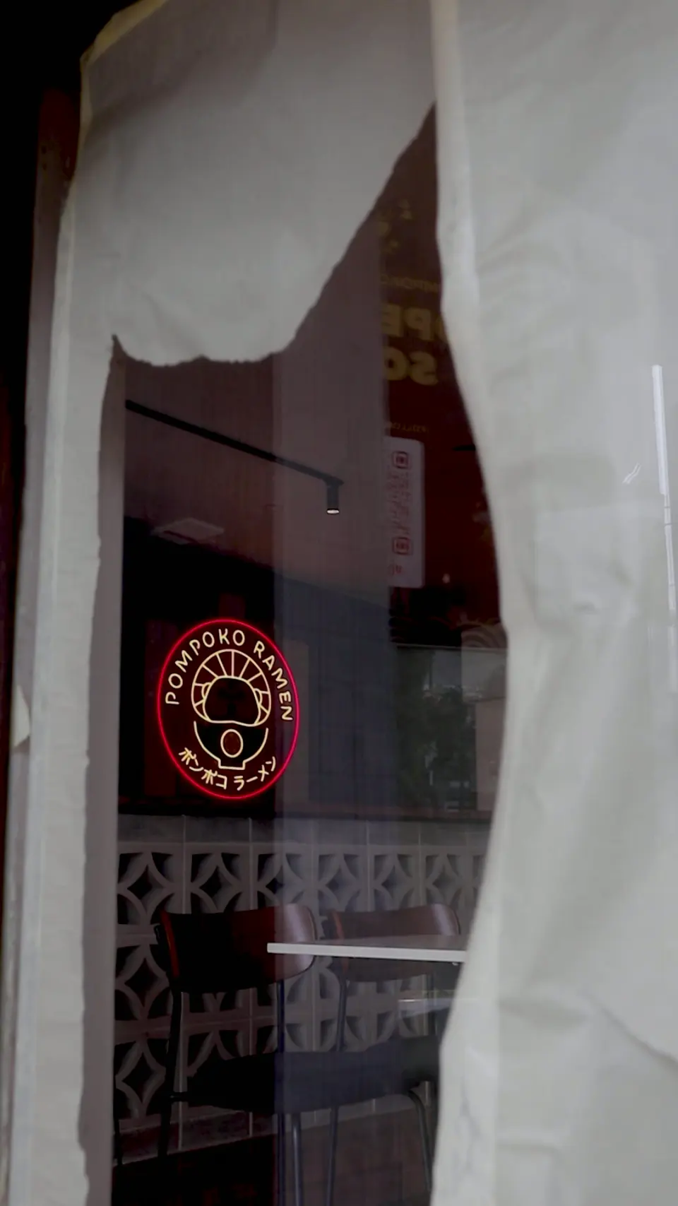 The neon Pompoko Ramen sign glowing through a curtained window