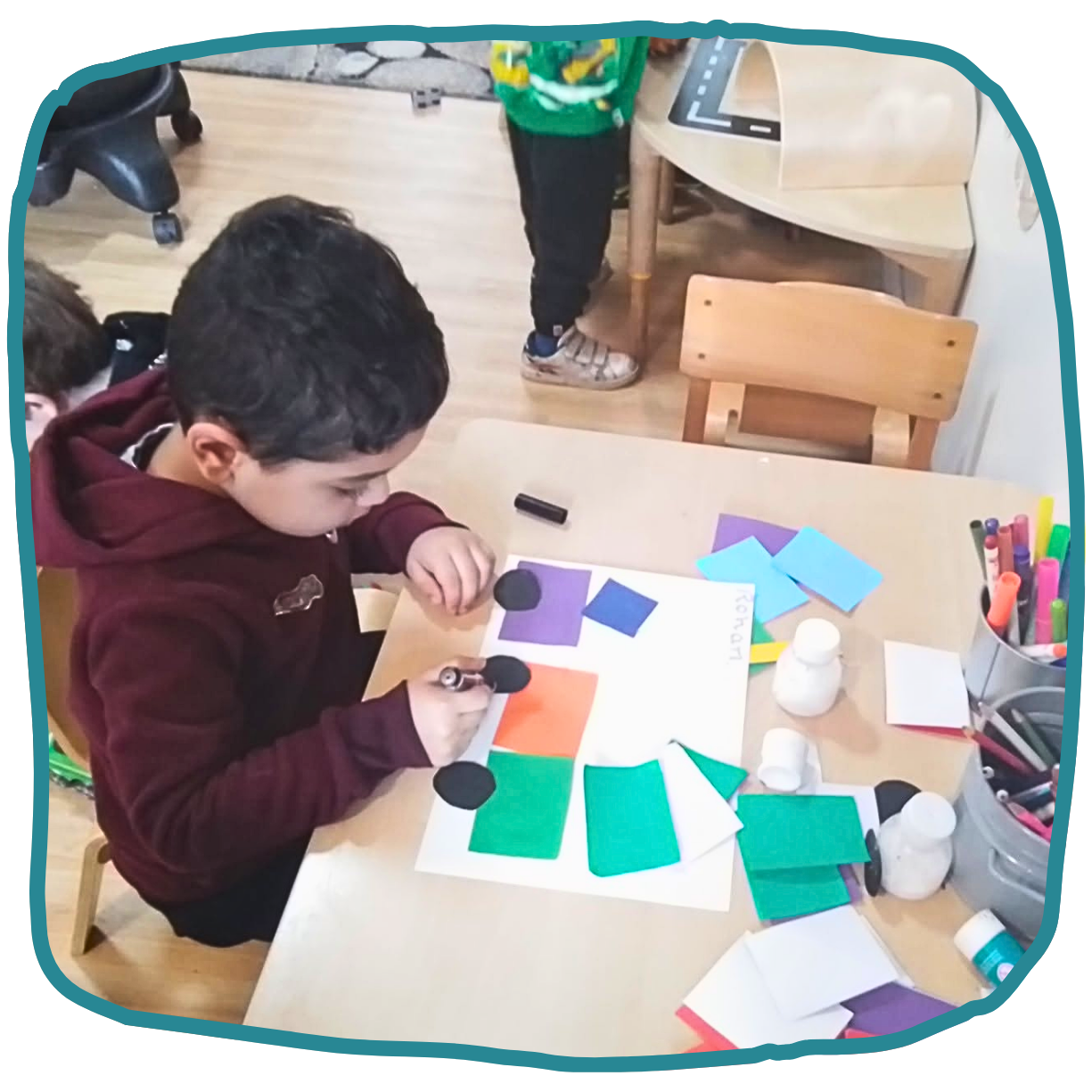A child arranges colored paper shapes on a worksheet at a classroom table, with markers, glue bottles, and more paper nearby at nursery in Merryfield