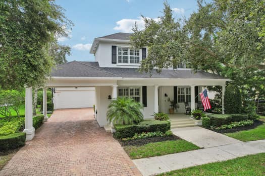 Two-story white Florida home with a brick paver driveway and wraparound porch under oak trees