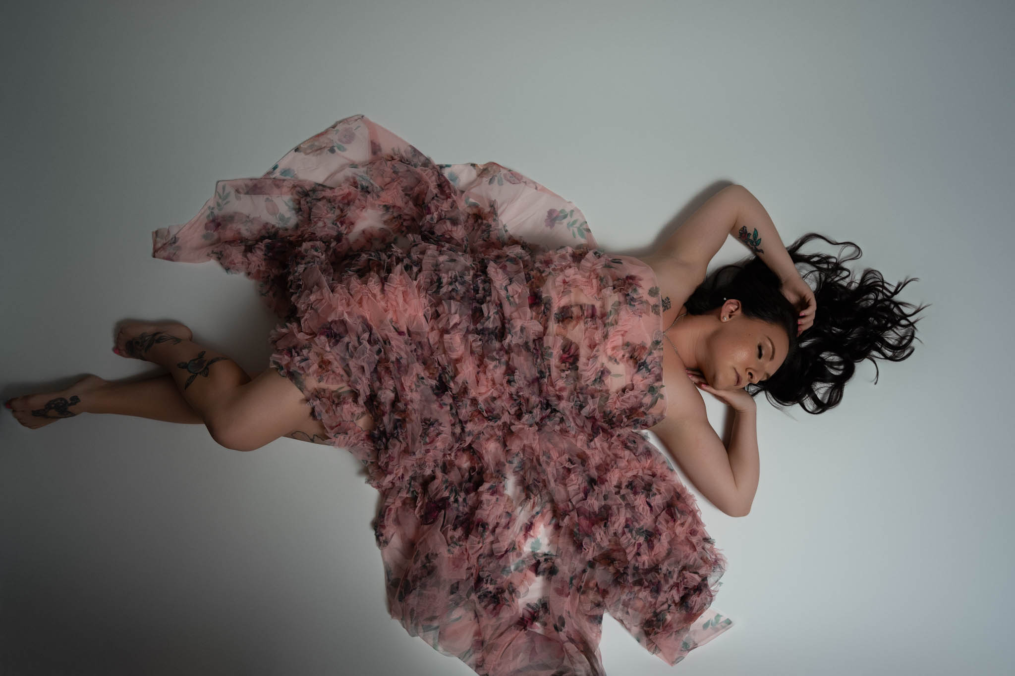 Artistic boudoir portrait of a tattooed woman lying gracefully in a flowing pink floral gown with dramatic movement and soft editorial lighting | Jessica Korff Studios Tucson, AZ Artistic boudoir portrait of a tattooed woman lying gracefully in a flowing pink floral gown with dramatic movement and soft editorial lighting | Jessica Korff Studios Tucson, AZ