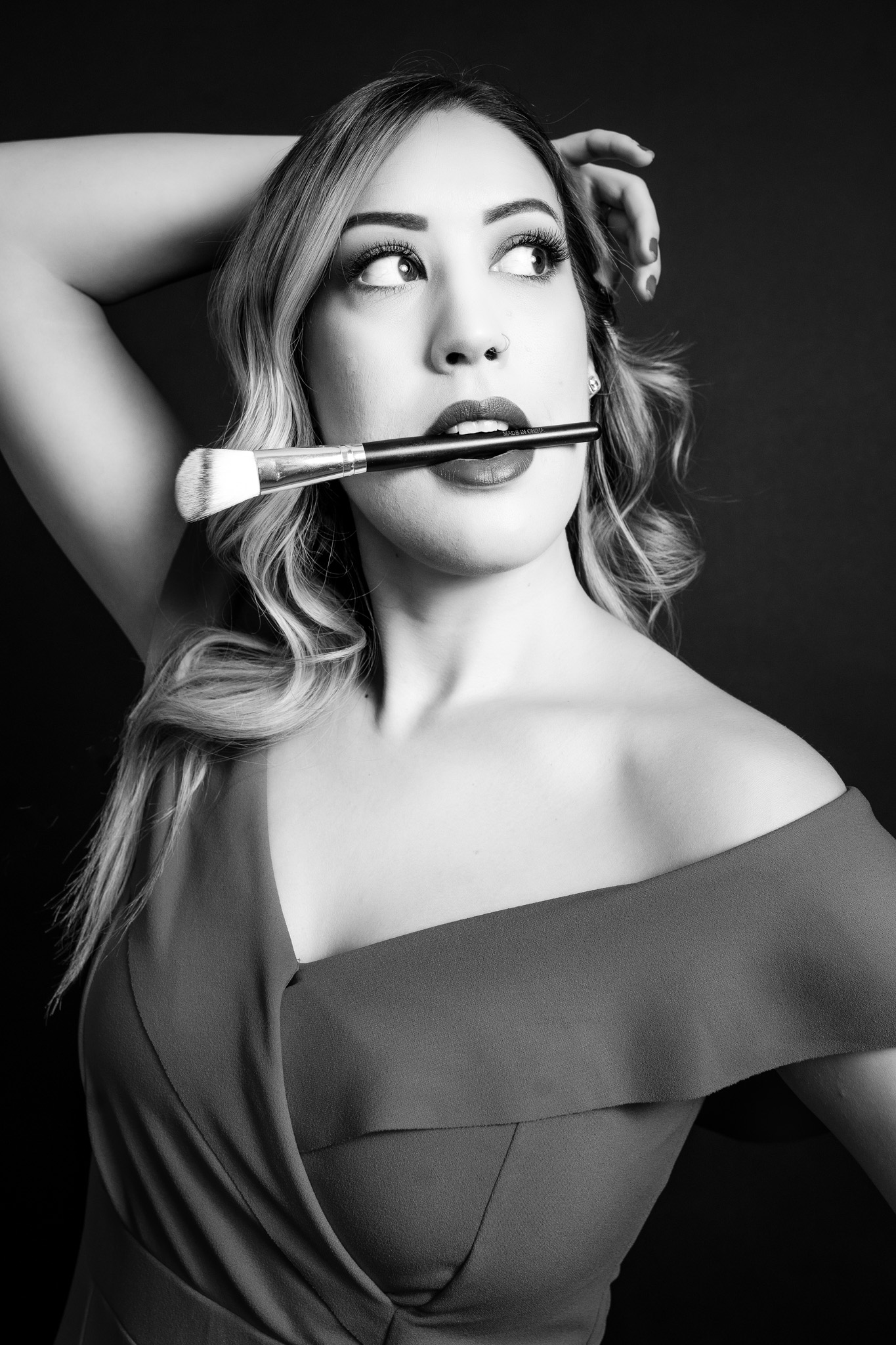 Black and white branding portrait of Tucson makeup artist holding a makeup brush between her lips while posing in an off-the-shoulder dress with glamorous makeup and curled hair | Jessica Korff Studios Tucson, AZ