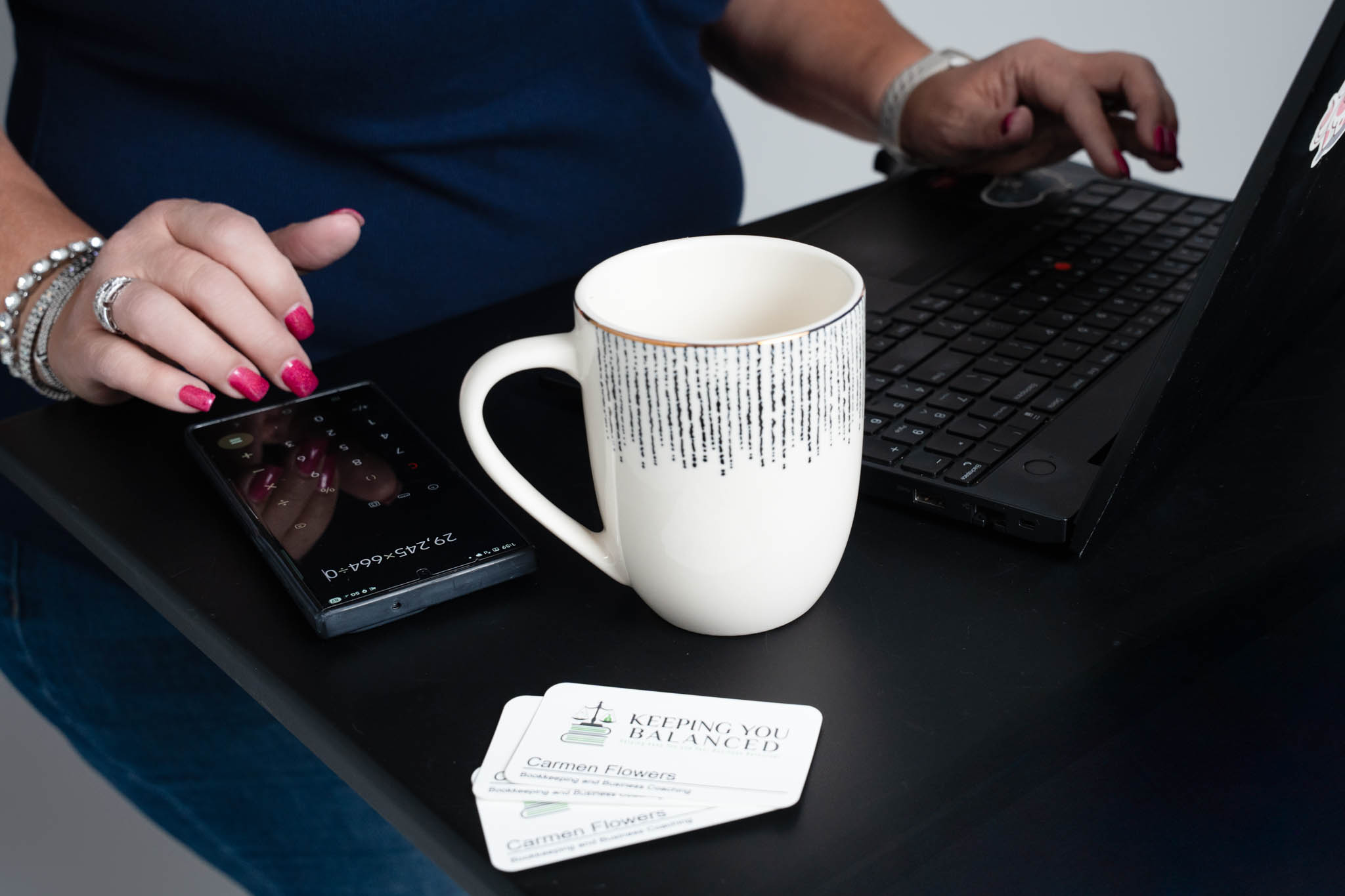 Close-up branding photo of a businesswoman working on a laptop beside a coffee mug, calculator app, and business cards for Keeping You Balanced bookkeeping services | Jessica Korff Studios Tucson, AZ