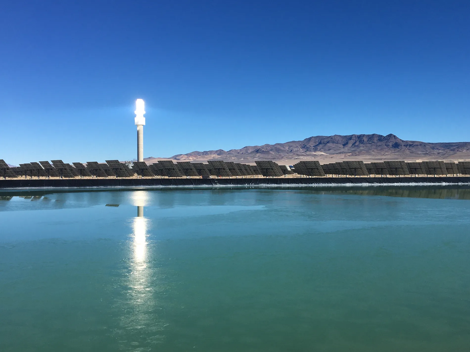 The lit receiver tower reflected in a still water basin at the edge of the solar field.