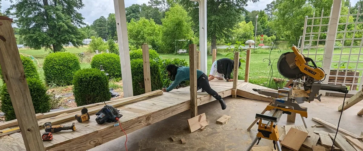 Workers building a wooden accessibility ramp on a back porch surrounded by greenery