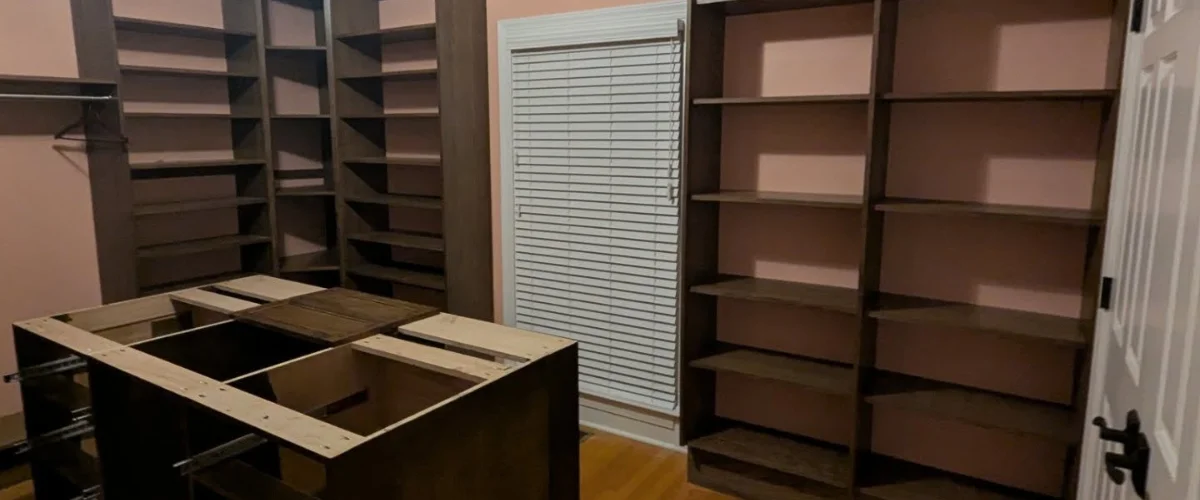 Custom walk-in closet shelving system under installation with multiple wooden storage units and window blinds.