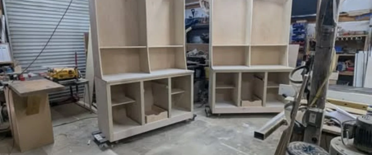 Two unfinished wooden cabinet units, with multiple shelves and compartments, standing in a workshop environment.