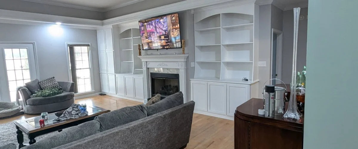A cozy living room with a fireplace, built-in white shelving, and comfortable gray furniture with large windows.