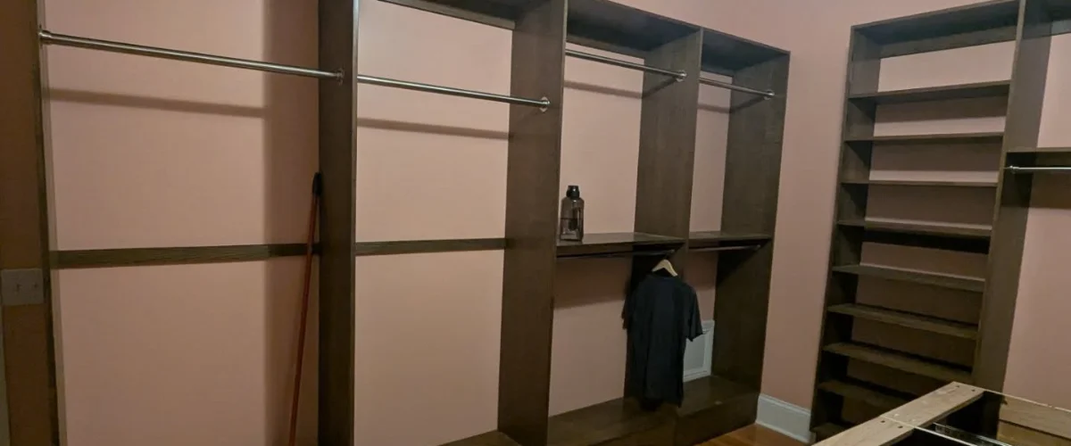 A wide view of a built-in closet with dark wood shelves and hanging rods against a light pink wall.