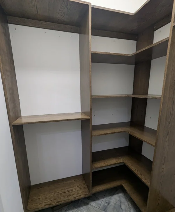An empty corner closet or pantry with dark brown finished wood shelves and white backing.