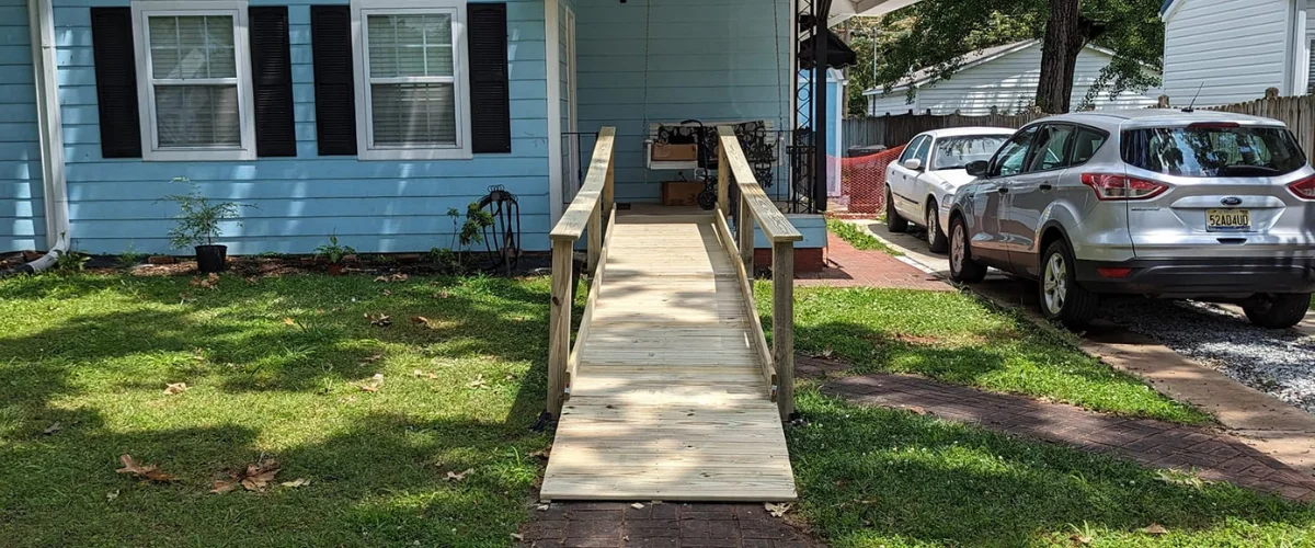 A wooden accessibility ramp leading to a light blue house with black shutters, with cars parked nearby. A wooden accessibility ramp leading to a light blue house with black shutters, with cars parked nearby.