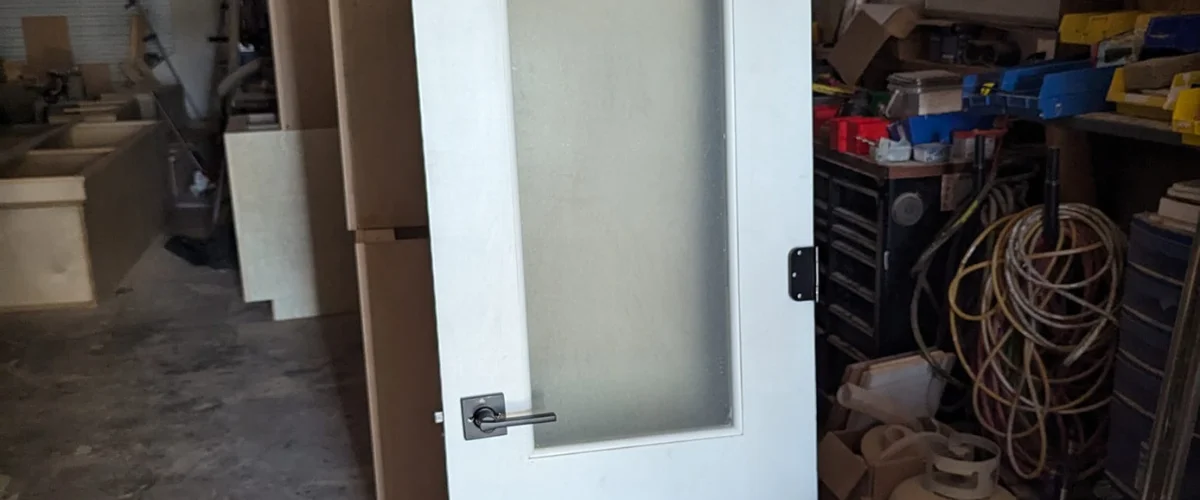 A close-up of a white interior door with a frosted glass panel and black handle, set in a messy workshop.