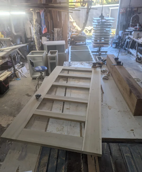 A light wood panel door in progress lies on a workbench in a dusty, sunlit woodworking shop.