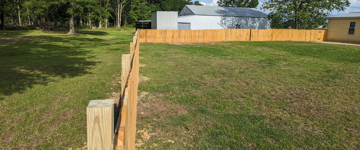 A new wooden fence runs through a grassy yard toward a white barn and distant trees under a blue sky.