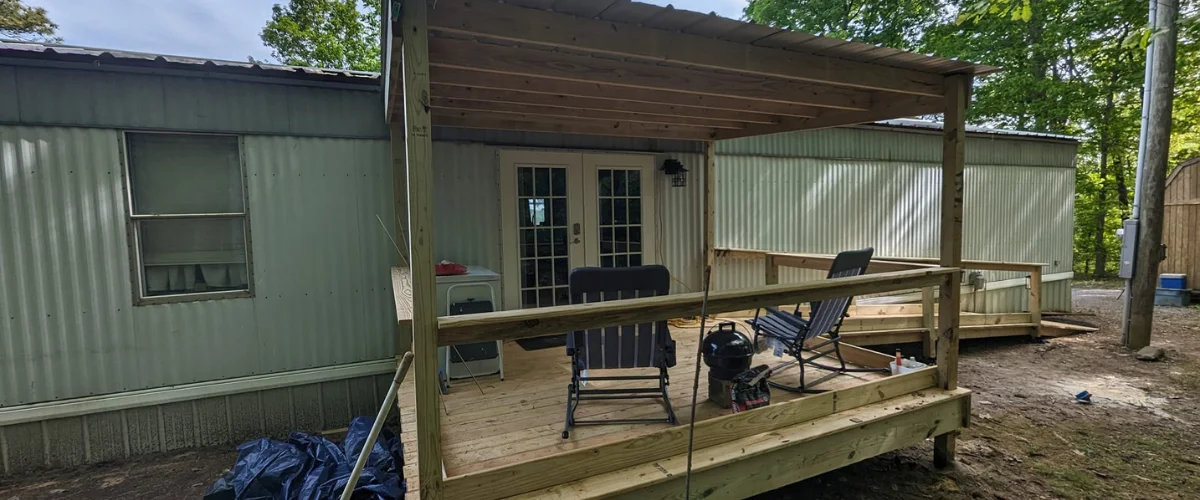 A newly built wooden deck with a covered patio and two chairs in front of a light green mobile home.