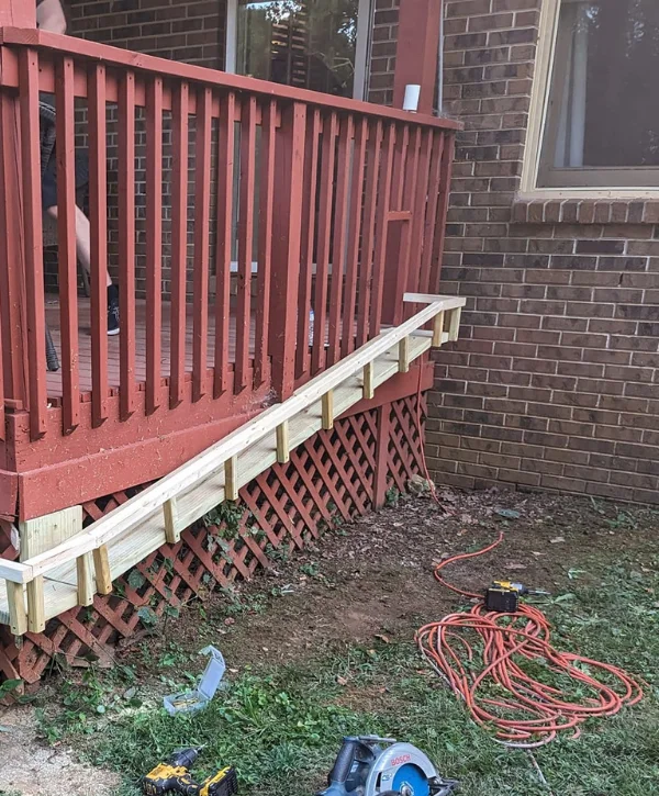 A partially built wooden ramp with new light-colored wood, attached to a red painted deck railing.