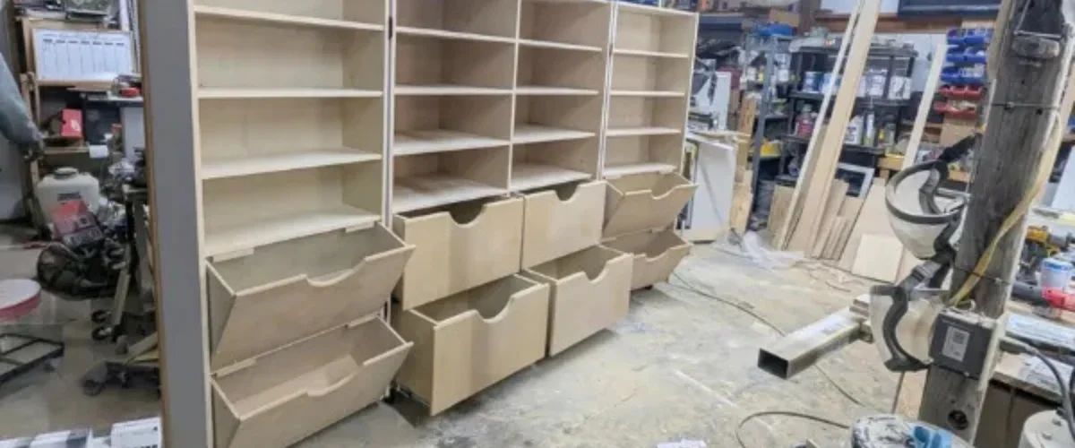 A close-up view of unfinished light wood shelving units with pull-out bins in a workshop environment. A close-up view of unfinished light wood shelving units with pull-out bins in a workshop environment.