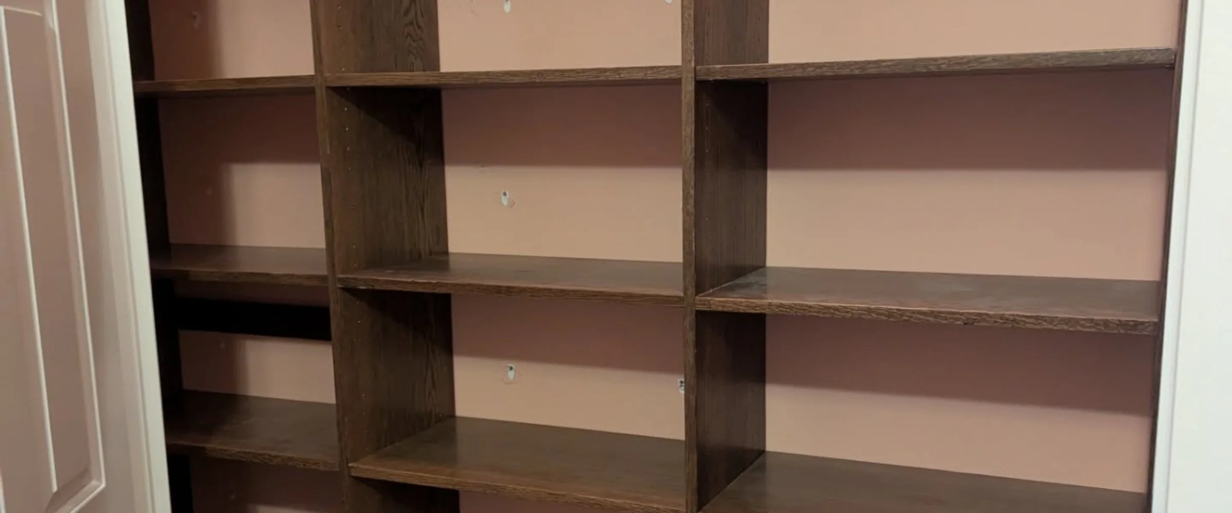 An empty, dark brown wooden shelving unit built into a closet or wall with a light pink back wall.