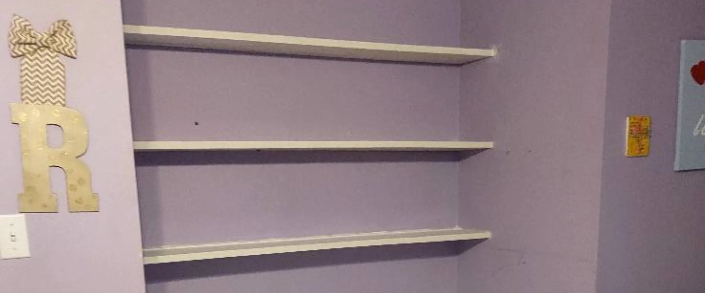 A room corner with light purple walls, featuring three empty white shelves and a large golden letter 'R' decor on the left. A room corner with light purple walls, featuring three empty white shelves and a large golden letter 'R' decor on the left.