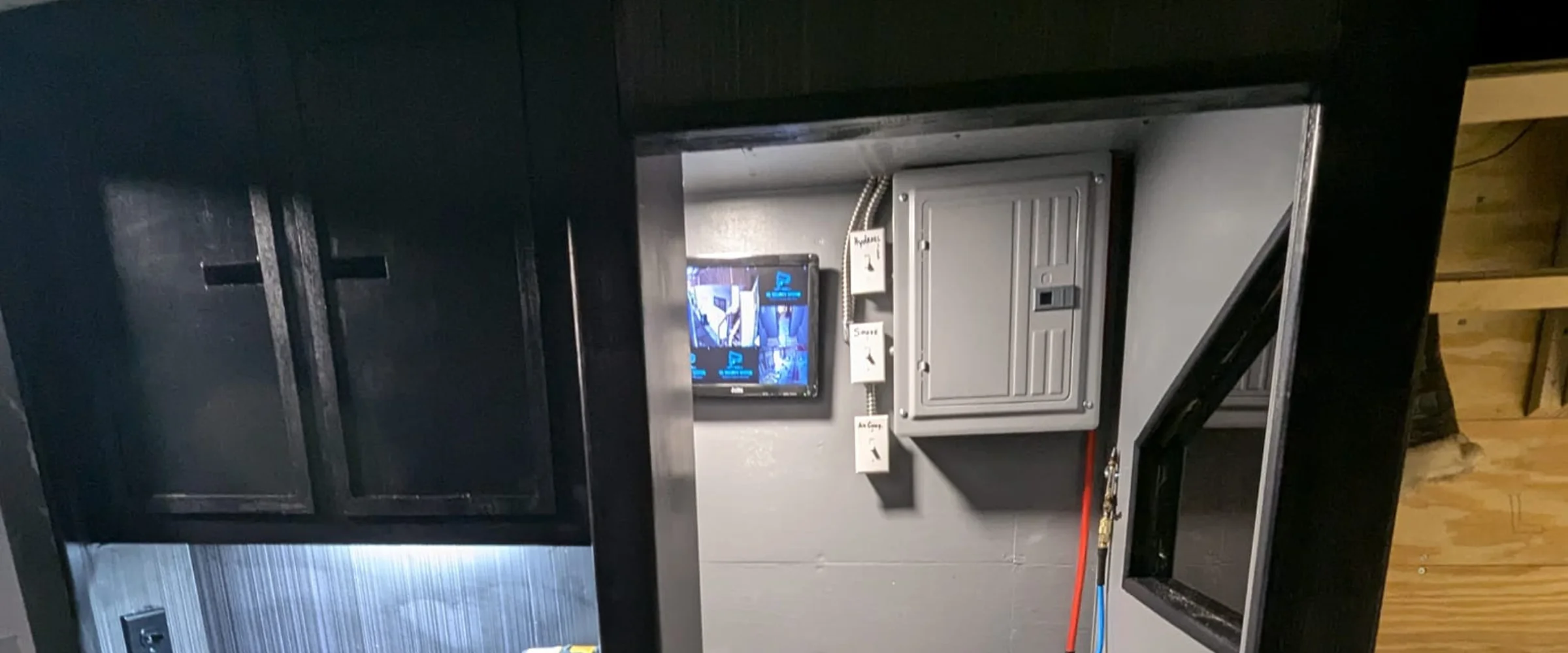 An interior view showing dark cabinets, a monitor displaying security feeds, a grey electrical panel, and switches. An interior view showing dark cabinets, a monitor displaying security feeds, a grey electrical panel, and switches.