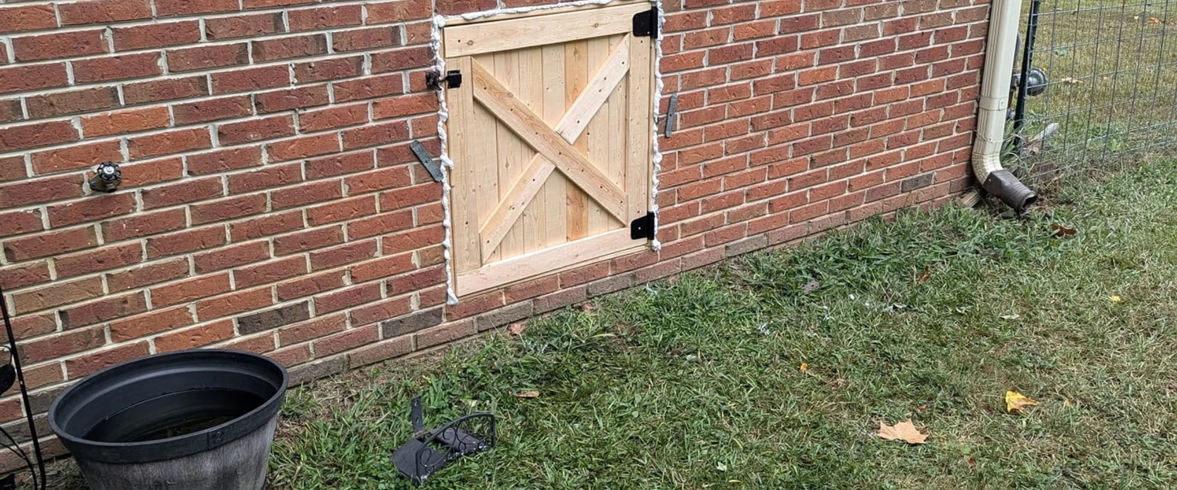A wooden X-brace access door on a red brick wall, next to a spigot and a downspout over green grass.