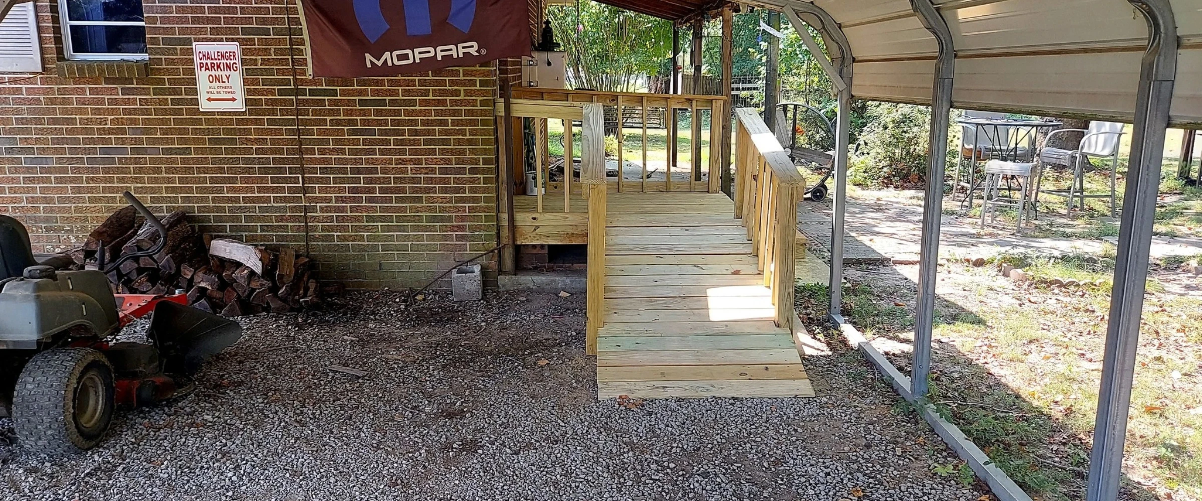A wooden ramp leading to a brick house with a Mopar flag and a 'Challenger Parking Only' sign, under a carport.
