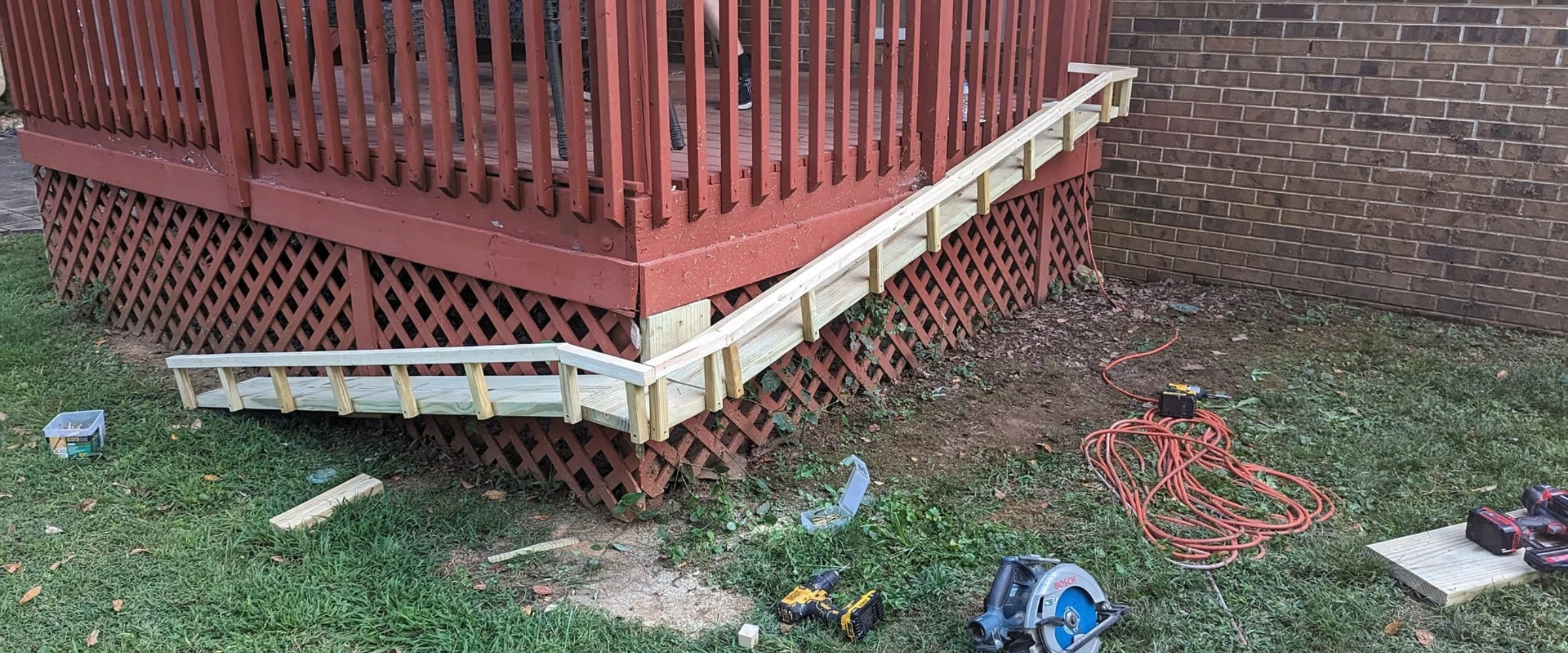 A newly built wooden wheelchair ramp leading up to a red deck with lattice skirting and tools on the ground.