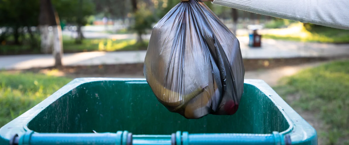 A person's hand in a white sleeve lowering a black trash bag into a green public bin outdoors.