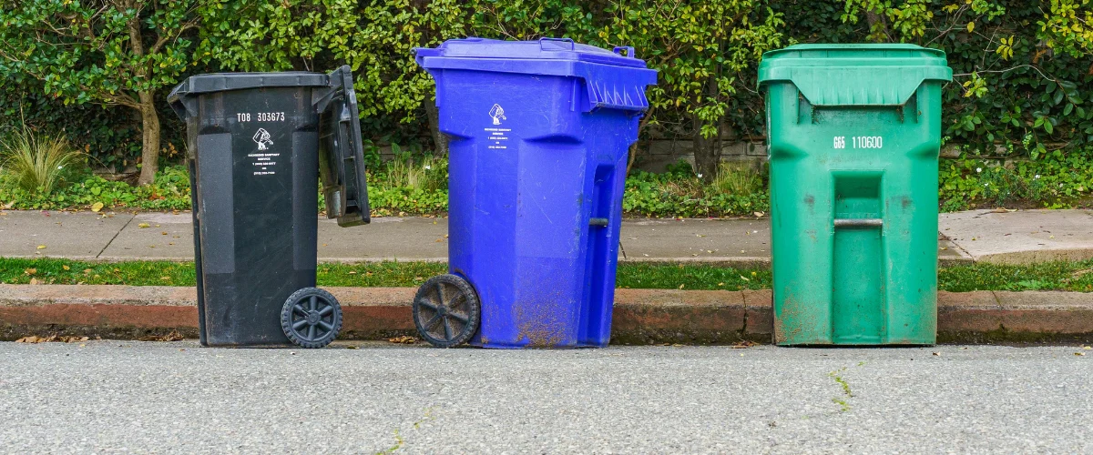Three industrial-sized recycling bins, black, blue, and green, stand in a row on the side of a street.