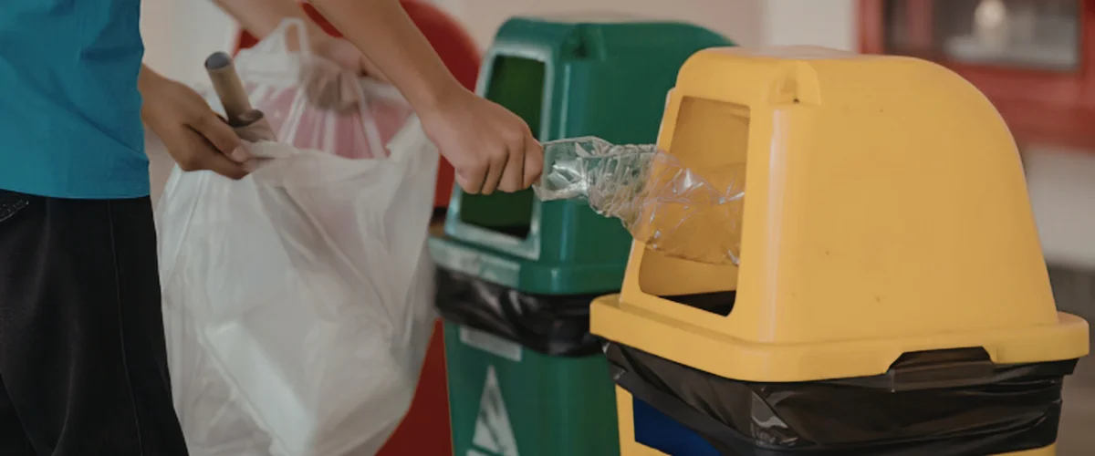 A person's hands placing a crumpled plastic bottle into a yellow recycling bin near a green bin.
