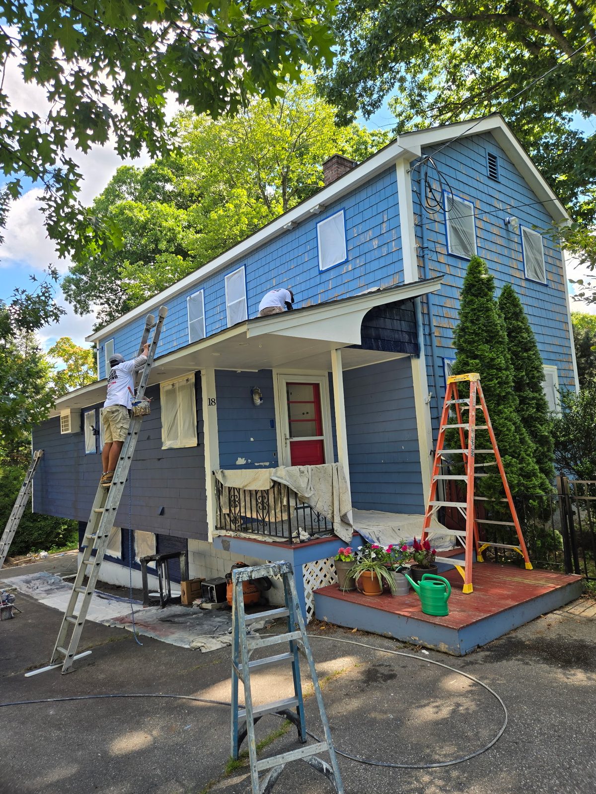 A two-storey house in faded light blue with primer patches, mid-prep, ladders against the siding.
