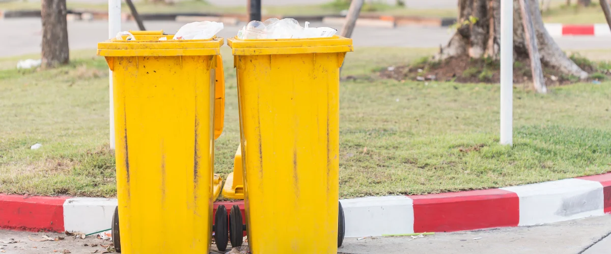 Two dirty yellow wheelie bins, with overflowing plastic recycling, sit on a concrete path beside a grass verge. Two dirty yellow wheelie bins, with overflowing plastic recycling, sit on a concrete path beside a grass verge.