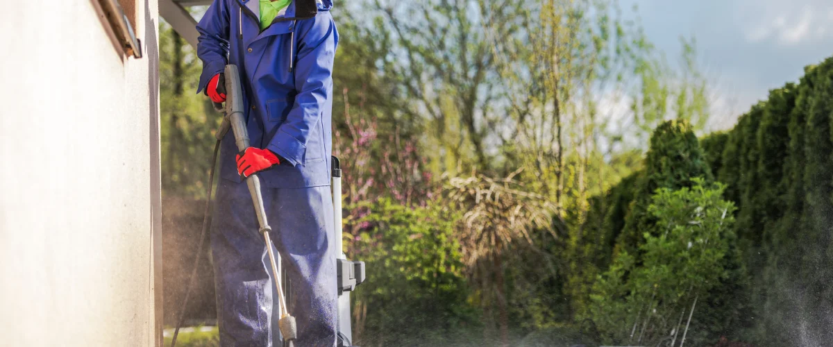 A person in a blue rain suit and red gloves pressure washing an outdoor wall during the daytime. A person in a blue rain suit and red gloves pressure washing an outdoor wall during the daytime.