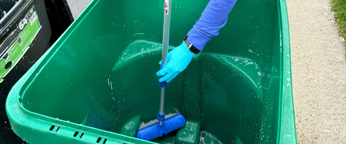 A person in blue gloves and sleeve cleaning a large green trash can with a blue scrub brush. A person in blue gloves and sleeve cleaning a large green trash can with a blue scrub brush.