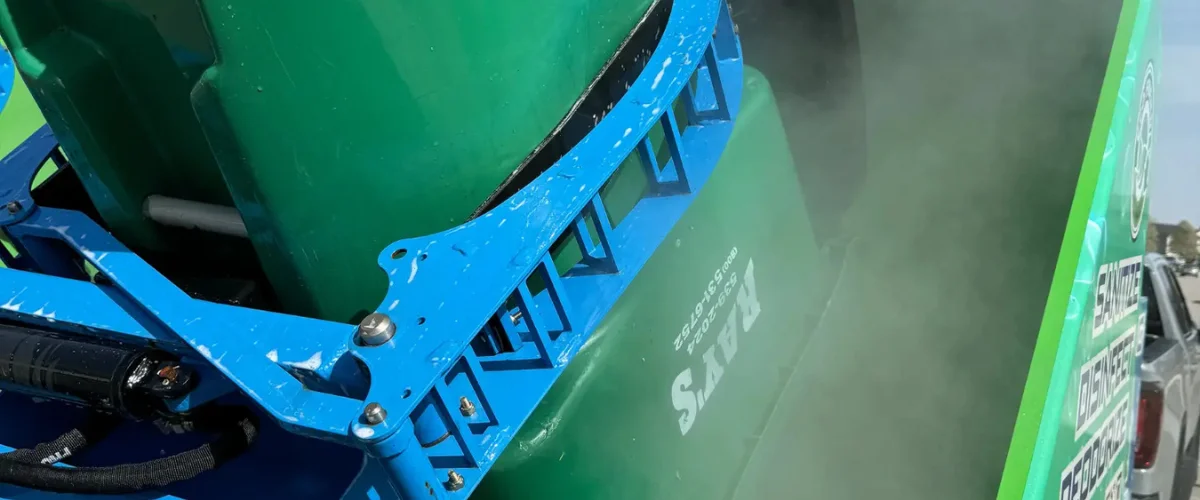 A close-up of a green sanitizing machine with blue hydraulic arms emitting a fine mist, parked on a sunny day.