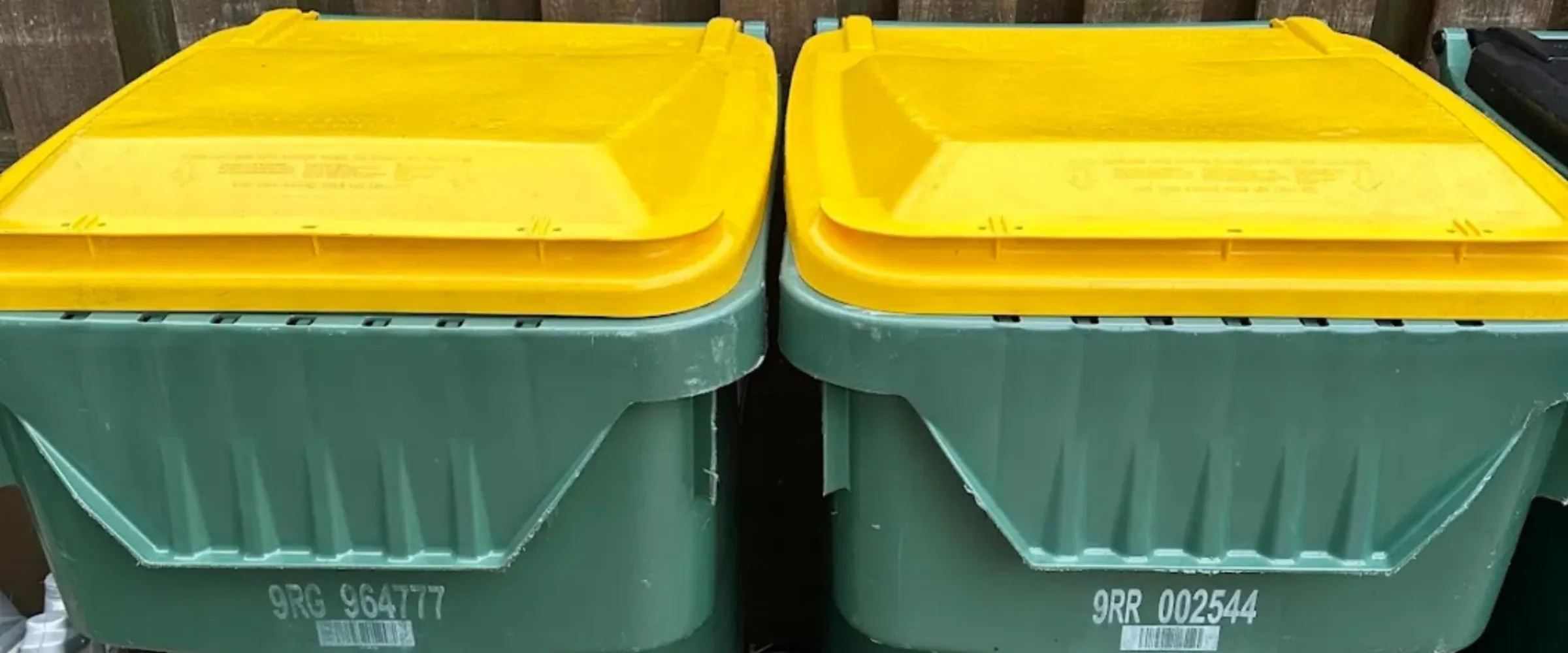 Two green recycling bins with bright yellow lids stand side-by-side, displaying barcodes and identification numbers.